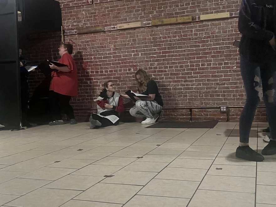 The Firebringer cast crouched and seated along the studio wall during rehearsal