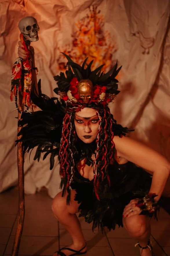 Molag crouches in a black feathered costume and flame headdress beside a skull-topped staff in Firebringer