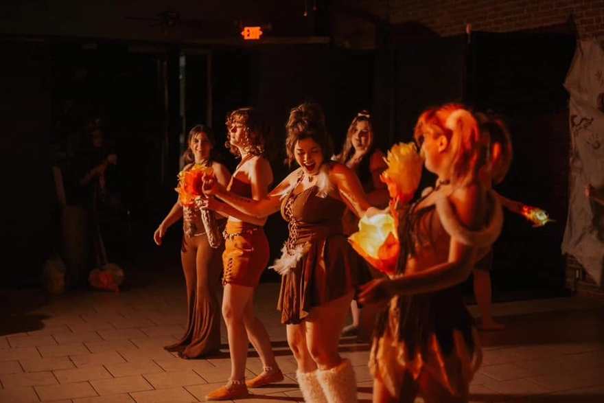 The Firebringer tribe in costume gathers on stage, one member holding a glowing flame