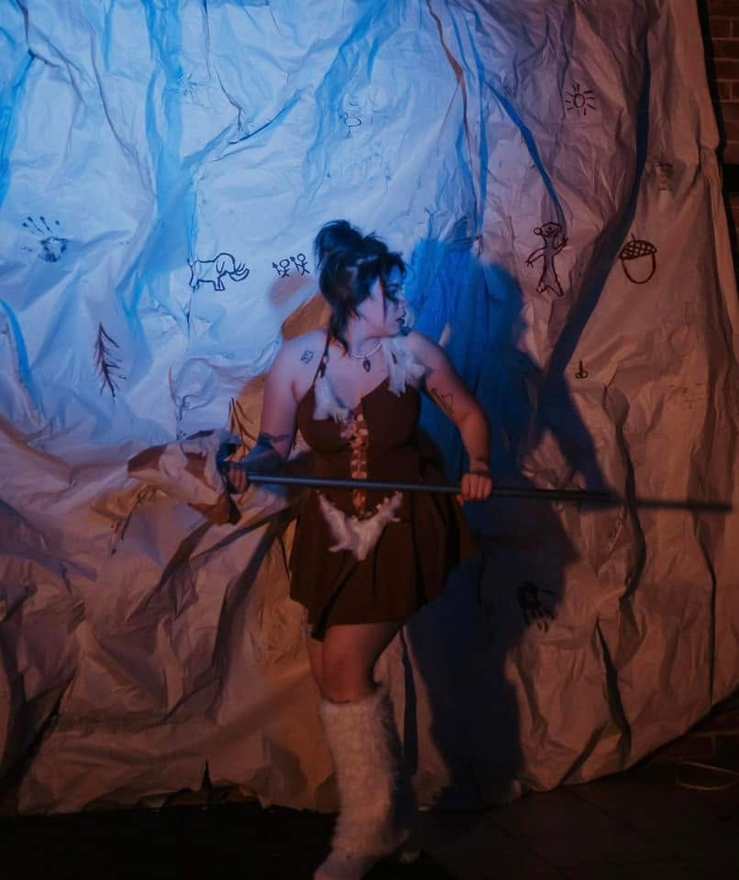 A Firebringer performer grips a spear before the blue-lit cave wall and its cave paintings